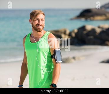 Mann mit Tanktop, Uhr, Smartphone-Armband, kabelgebundenen Ohrhörern, die am Strand laufen, Kopierraum Stockfoto