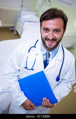 Männlicher Arzt im Patientenzimmer mit blauem Aktenordner mit Stethoskop neben dem Krankenhausbett Stockfoto