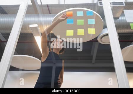 Asiatische Geschäftsfrau, die bunte Haftnotizen auf Glaswand in industriellen Büroflächen platziert Stockfoto