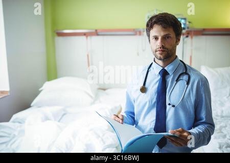 Männlicher Arzt, der neben dem Krankenhausbett steht und die Patientenakte mit Stethoskop überprüft, Kopierraum Stockfoto