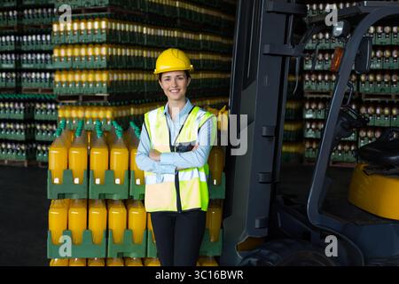 Frau mit Sicherheitsweste und Schutzhelm, die Getränkepaletten im Lager neben dem Gabelstapler inspiziert Stockfoto