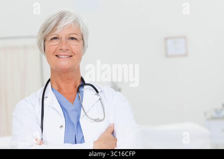 Leitende Ärztin, die lächelnd im Untersuchungsraum steht und eine Stethoskop-Brille trägt Stockfoto