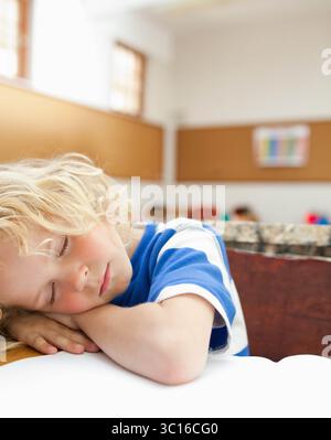 Schuljunge, der mit leerem Notizbuch-Papier Kopf auf gefalteten Armen auf einem hölzernen Schreibtisch im Klassenzimmer ruht Stockfoto