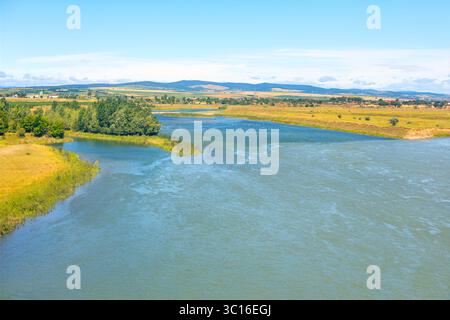 Ruhiger Wasserweg, der die Weite der umliegenden Landschaft widerspiegelt. Ein ruhiger Fluss fließt durch die grasbewachsene Ebene, die von Vegetation gesäumt ist Stockfoto