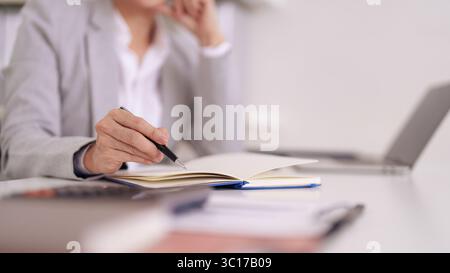 Nahaufnahme eines Erwachsenen, der in ein blaues Notizbuch schreibt. Ein Laptop sitzt in professioneller Büroumgebung auf dem Schreibtisch. Ideal für Produktivitätsthemen, Bu Stockfoto