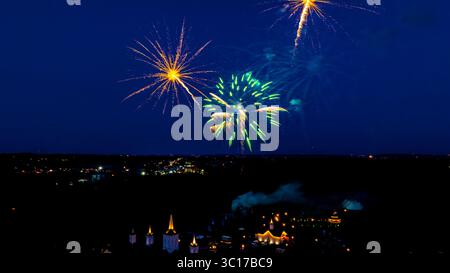 Ein helles Feuerwerk in verschiedenen Farben platzt nachts über einer Stadtlandschaft. Darunter sind Gebäude und Bäume zu sehen, die die festliche Atmosphäre dieser Sommerfeier noch verstärken. Stockfoto
