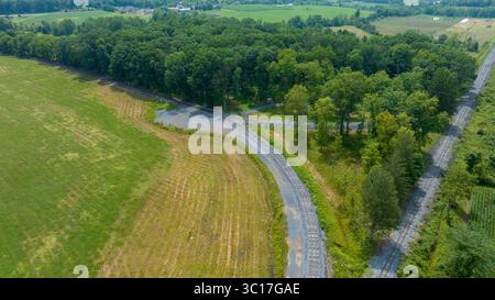 Dieser malerische Blick fängt eine gewundene Straße ein, die von lebhaftem Grün und offenen Feldern umgeben ist, und zeigt die Schönheit des ländlichen Lebens im Sommer. Die üppigen Bäume und Felder schaffen eine ruhige Atmosphäre. Stockfoto