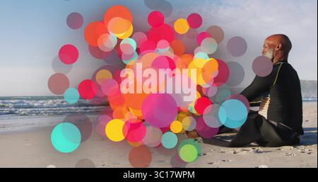 Sitzender älterer Mann im Taucheranzug, der auf den Ozean am Sandstrand blickt, mit bunten Bokeh, Kopierraum Stockfoto