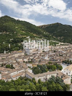Blick aus der Vogelperspektive auf eine historische Stadt, eingebettet unter einem grünen Berg, wo Terrakotta-Dächer im Kontrast zu den hellen Steinen von alten Gebäuden stehen, Gubbio, Provinz Perugia, Italien. Stockfoto