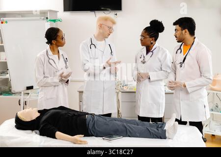 Vier Medizinstudenten, die Handschuhe und weiße Mäntel tragen, üben während eines Trainings die HLW an einer medizinischen Attrappe Stockfoto