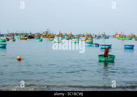 Mui ne, Vietnam, 22. März 2025: Ein Fischer rudert ein traditionelles Rundkorbboot durch ein Meer farbenfroher Fischereifahrzeuge Stockfoto