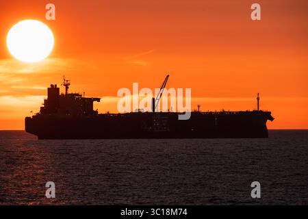 Silhouette eines großen Frachtschiffes, Öltanker, der auf dem Meer segelt, dramatischer orangefarbener Sonnenuntergang, Sonne über dem Horizont. Stockfoto
