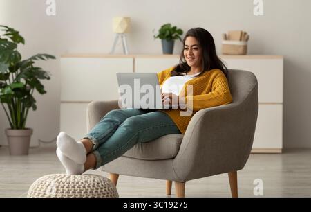 Aufgeregte arabische Frau in legerer Kleidung, die von zu Hause aus online arbeitet, in einem bequemen Sessel sitzt und einen Laptop benutzt, um Platz zum Kopieren zu nutzen Stockfoto