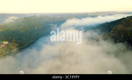 Aus der Vogelperspektive auf die nebeligen Hügel, die einen ruhigen Fluss umgeben. Eine friedliche Luftaufnahme des Nebels, der über grüne Hügel gleitet, und ein ruhiger Fluss, der sich durch die schlängelt Stockfoto