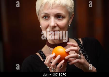 Eine Frau mit kurzen, blonden Haaren hält eine leuchtende orange Tomate in den Händen. Sie trägt ein schwarzes Kleid mit Spitzendetails und ein dunkles Burgunderrot Stockfoto