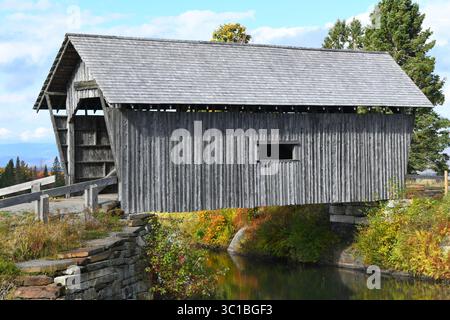 Das Äußere der A. M. Foster Covered Bridge in Cabot, Vermont, ist verwittert und verblasst. Es hat ein kleines Fenster und erstreckt sich über einen Teich. Stockfoto