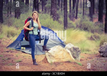 Weibliche Wanderer, die in der Kiefernwaldlichtung vor einem zwei-Personen-Kuppelzelt mit Rucksackgurt steht Stockfoto