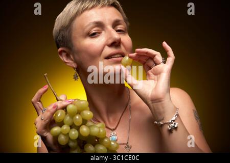 Eine Frau mit kurzen blonden Haaren hält einen Haufen grüner Trauben in den Händen. Sie trägt silberne Ohrringe und ein silbernes Armband. Sie ist auch getragen Stockfoto