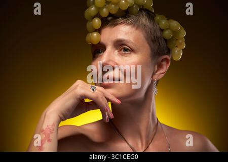 Eine Frau mit kurzen, hellbraunen Haaren trägt eine Krone aus grünen Trauben auf dem Kopf. Sie hat ein dezentes Tattoo am linken Arm und einen Ring am Finger. Stockfoto