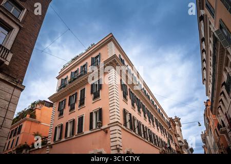 Flacher Blickwinkel auf ein pastellfarbenes, römisches Apartmentgebäude mit Fensterläden, Dachbegrünung und Quoins, die sich zwischen ihnen scharf erheben Stockfoto