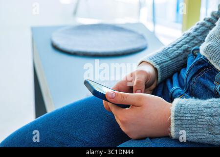 Nahaufnahme eines jungen Menschen in einem grauen Strickpullover und einer blauen Jeans mit Smartphone, während er auf einer Bank in einem hellen Innenraum sitzt. Stockfoto