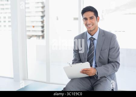 Geschäftsmann aus dem Nahen Osten, der in der Lobby des Büros sitzt und auf dem Klemmbrett an den Fenstern mit Uhr sitzt Stockfoto