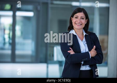Reife Geschäftsfrau im Anzug, die in den Glastüren der Lobby lächelt und die Arme ausstellt Stockfoto