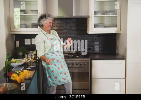 Ältere Frau mit Schürze, die tanzt, Paprika auf der Theke in der modernen Küche mit Salatschüssel hackt Stockfoto