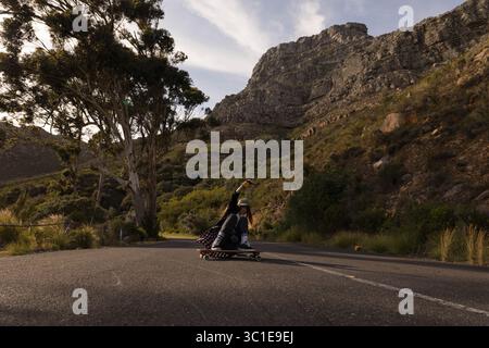 Weibliche Skateboarderin, die tief auf Longboard hockt und mit weißem Helm die Bergstraße hinuntergleitet Stockfoto