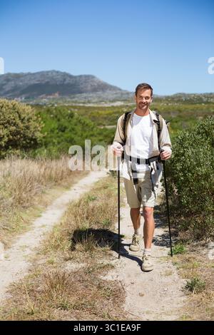 Man wandert auf einem gewundenen Feldweg durch grasbewachsenes Buschland mit Trekkingstöcken und Rucksack Stockfoto