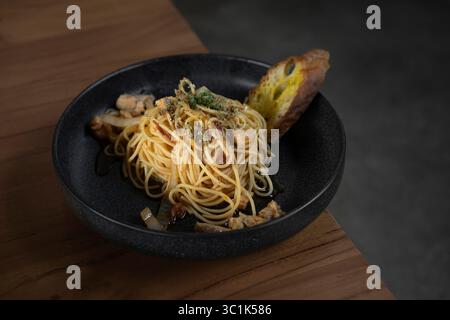 Spaghetti-Pasta mit Hühnchen und Knoblauchbrot in einer schwarzen Schüssel auf einem Holztisch. Italienisches Gourmetgericht in Restaurant-Atmosphäre. Schließen Sie das Essen. Stockfoto