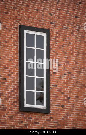 Ein hohes klassisches weißes Fenster mit schwarzem Rahmen in einer roten Ziegelwand. Minimalistisches architektonisches Detail einer Gebäudefassade mit einem klaren Muster. Stockfoto