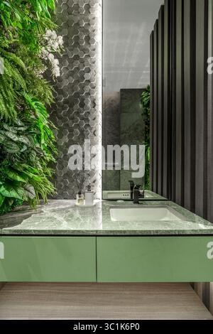 Moderner Kosmetikbereich im Bad mit grünem Schrank, grauer Marmorplatte und einem üppigen vertikalen Garten. Biophile Inneneinrichtung mit sechseckigen Fliesen und Spiegel. Stockfoto