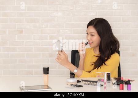 Die lächelnde asiatische Frau nimmt ein Schönheitslehrbuch auf, indem sie Lippenstift vor einer Kamera mit verschiedenen Make-up-Produkten auf einem weißen Schreibtisch aufträgt. Stockfoto