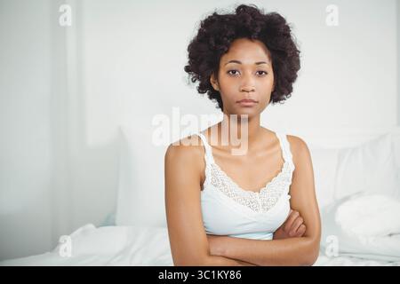 Afroamerikanerin sitzt und kreuzt die Arme, während sie auf weißem Bett mit Kissen in die Kamera blickt Stockfoto