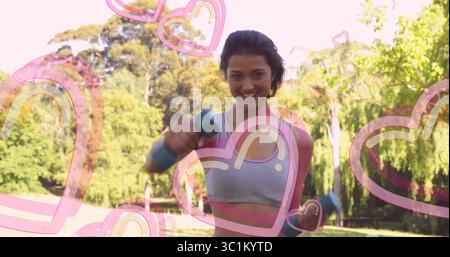 Zwei blaue Kurzhanteln halten, asiatische Frau in Sportbekleidung, die im Park trainiert, mit rosa Herzüberzügen Stockfoto