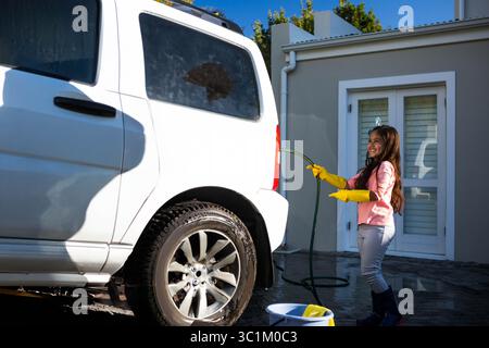 Gartenschlauch, der Wasser auf weißen SUV in Vorstadteinfahrt mit gelben Handschuhen und blauem Eimer sprüht Stockfoto