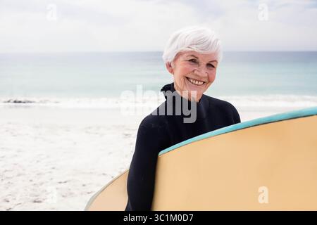 Senior Surfer mit gelbem Surfbrett am Sandstrand und Blick auf das türkisfarbene Meer unter bewölktem Himmel Stockfoto