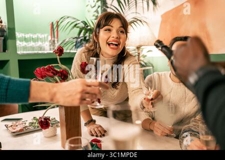 Eine schöne junge Frau mit Pony lacht vor Freude, während sie ihr Glas hochhebt, um mit einer Gruppe von Freunden bei einem fröhlichen weihnachtsabend anzustoßen Stockfoto