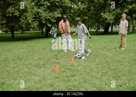 Multiethnische Kinder und männlicher Lehrer spielen gemeinsam Fußball im Park Outdoor Stockfoto