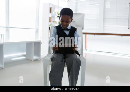 Afroamerikanischer Junge in formeller Kleidung, der auf weißem Stuhl in einem modernen Zimmer sitzt und ein Tablet hält Stockfoto