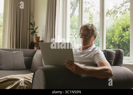 Älterer Mann sitzt auf grauem Sofa im Wohnzimmer mit Laptop neben Topfpflanzen Stockfoto