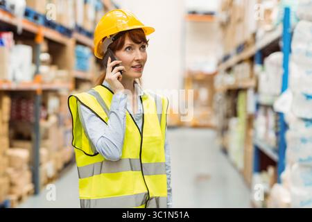 MittelErwachsene Frau mit Sicherheitsweste und gelbem Schutzhelm, die auf dem Smartphone spricht und das Lager inspiziert Stockfoto