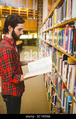 Erwachsener Mann, der im Büchergang zwischen hölzernen Bücherregalen steht, während er offenes Buch liest, Kopierraum Stockfoto