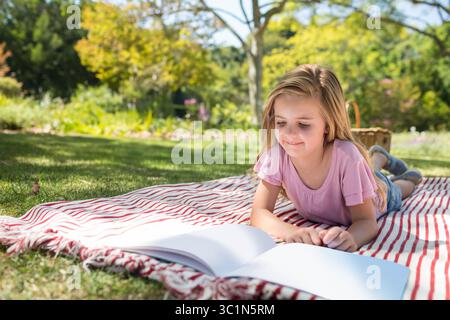 Weibliches Kind, das auf einer Picknickdecke liegt und ein offenes Buch in der Nähe des Korbs im Park liest, Kopierraum Stockfoto