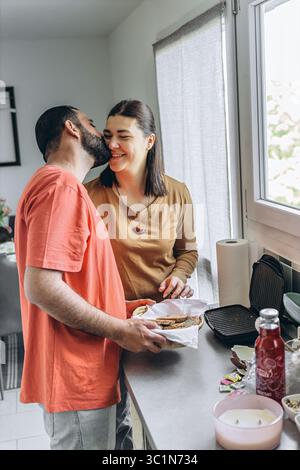Ein liebevolles Paar teilt einen zärtlichen Moment in der Küche, während sie zusammen Frühstück vorbereiten. Der Mann küsst die Frau auf die Wange, während sie lächelt, Holdi Stockfoto