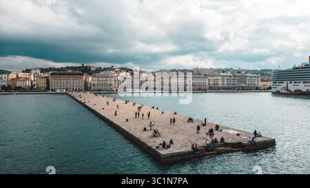 Aus der Vogelperspektive auf einen langen Pier, der sich in die Adria erstreckt, wo sich Menschen vor dem Hintergrund historischer Gebäude und eines Kreuzfahrtschiffs treffen, Triest, Friaul-Julisch Venetien, Italien. Stockfoto