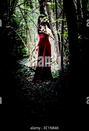 Eine Frau in einem langen roten Kleid steht in einem dunklen Wald, teilweise verdeckt von Bäumen und Laub. Stockfoto