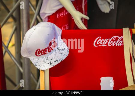 Stilvolle weiße Coca-Cola-Kappe mit Spritzermuster auf dem Stuhl mit rotem Branding während einer Veranstaltung im Freien. Coca-Cola-Begriff als Symbol wirklicher Magie, f Stockfoto