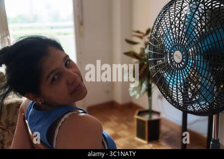Eine Frau sitzt in einem Zimmer mit reinem Ventilator. Sie trägt im Sommer ein Hemd mit blauem Ärmel Stockfoto